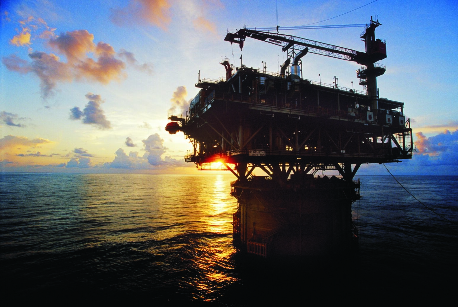 At dusk, the sun’s rays illuminate an offshore oil rig, casting a warm glow across the water.