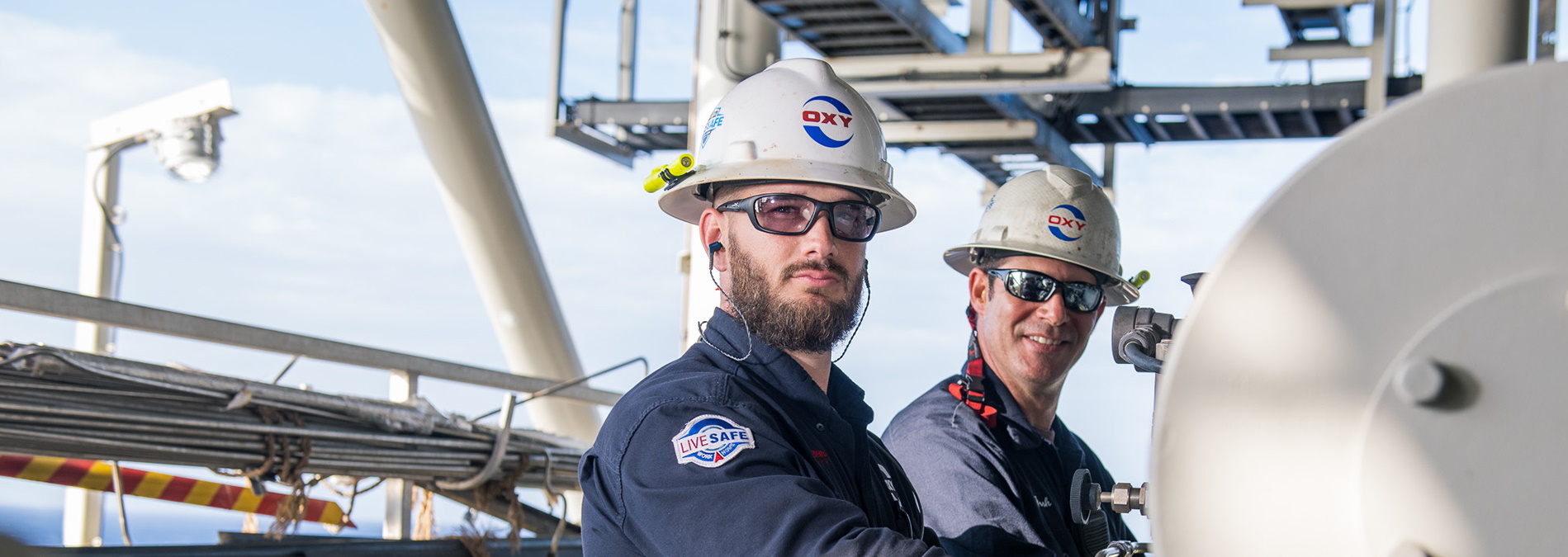 Two workers, working on a rig, wearing hard hats and smiling.